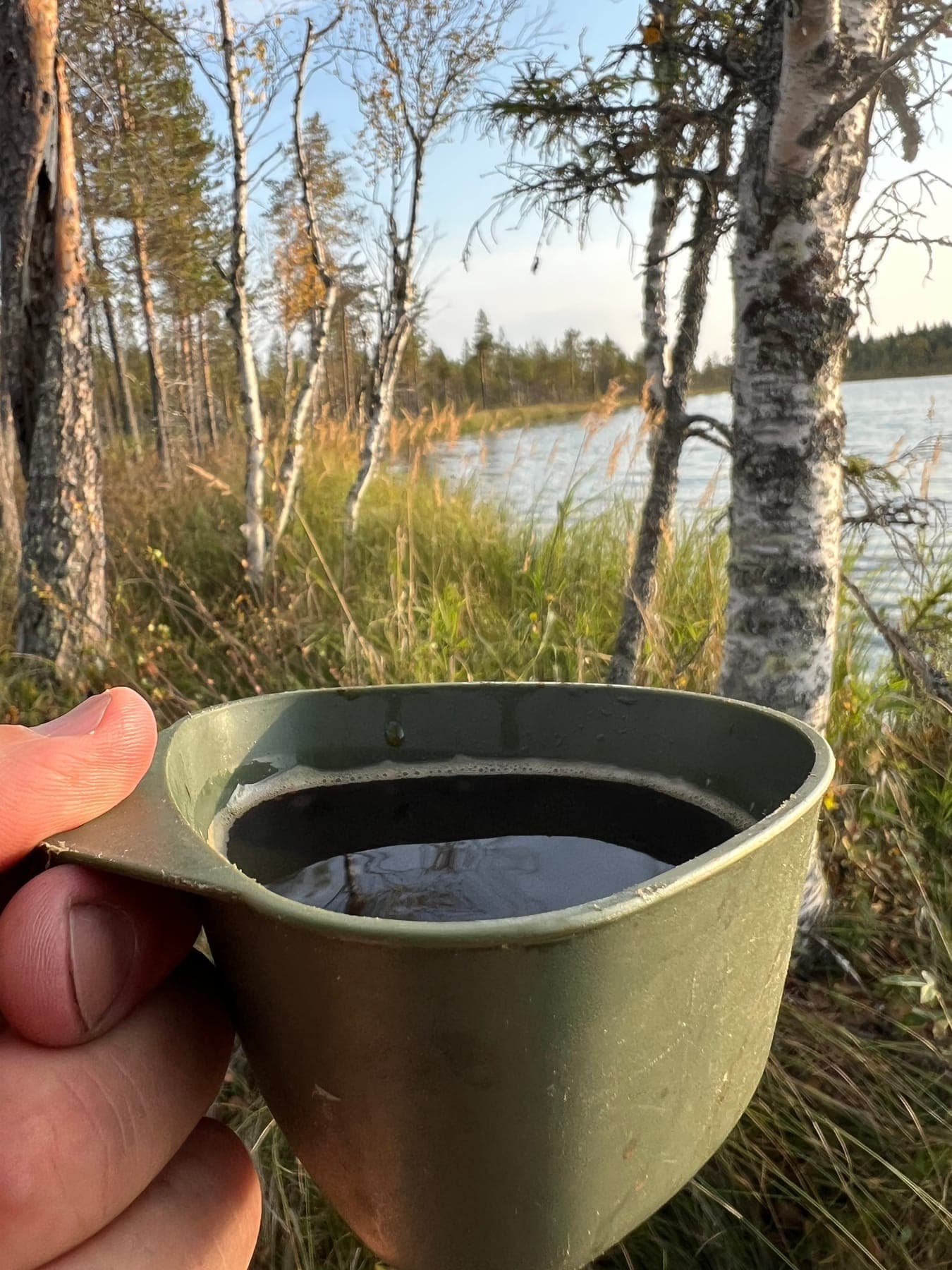 Hand håller en kåsa med kokkaffe vid sjöstrand i gyllene kvällssol